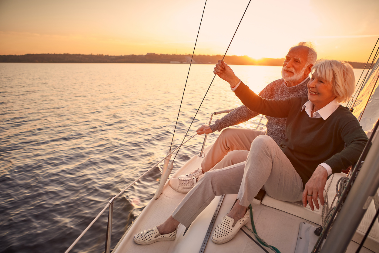 Enjoying luxury life. Beautiful happy senior couple in love relaxing on the side of sailboat or yacht deck floating in sea at sunset, looking at amazing evening view. Vacation, travelling, boat tour